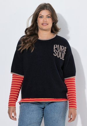 Vrouw met lang golvend haar, draag een zwarte trui met korte mouwen met de tekst "PURE SOUL" over een rood-oranje gestreept overhemd met lange mouwen en spijkerbroek.