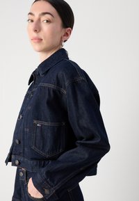 Mørk denim kort jakke med krage, frontlommer og knappelukking. Har kontrastfarget oransje søm. Glatt tekstur.