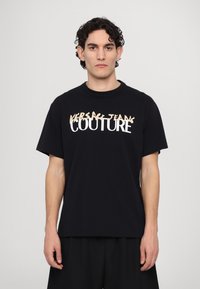 Juoda medvilninė t-shirta su trumpomis rankovėmis, su ryškiu baltu ir šviesiai smėlio spalvos tekstu "VERSACE JEANS COUTURE" mišriame šriftų stiliuje.