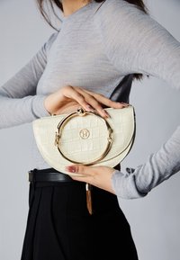 Borsa crema con motivo coccodrillo, dotata di un manico rotondo dorato, tenuta in mano, abbinata a una maglietta grigia a maniche lunghe e pantaloni neri.