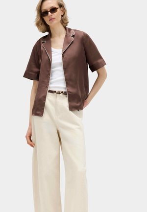 Kvinde iført brun satinskjorte med korte knapper, hvid tanktop, beige bukser med vide ben, solbriller og et brunt bælte.