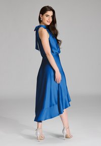 Niebieska satynowa sukienka z kokardą na szyi, bez rękawów, zwiewny dół i warstwowy brzeg, połączona ze srebrnymi szpilkami.