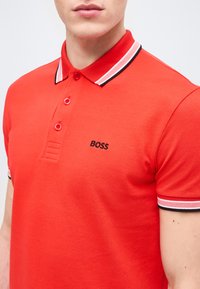 Homme portant un polo rouge vif avec un col et des manches rayés noirs et blancs, et un logo "BOSS" noir sur le côté gauche de la poitrine.
