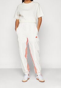 Henkilö, jolla on päällään valkoiset cargo-housut oransseilla raidoilla ja pelkkä valkoinen t-paita, seisoo kädet taskuissa tasaisella taustalla.