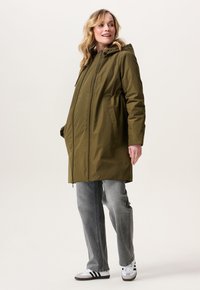Vrouw in een olijfgroene winterjas met capuchon over grijze jeans en witte sneakers, staand en met een glimlach naar de zijkant kijkend.