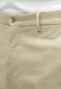 Μπεζ chinos από απαλό ύφασμα; διαθέτει επίπεδο μπροστινό μέρος, κλείσιμο με ένα κουμπί και διακριτικές λεπτομέρειες στις πλευρικές τσέπες.