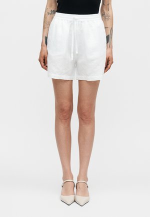 Personne portant un short blanc à cordon de serrage et des talons beiges à bout pointu, avec des tatouages visibles sur les avant-bras sur un fond uni.