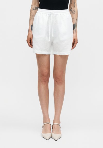 Personne portant un short blanc à cordon de serrage et des talons beiges à bout pointu, avec des tatouages visibles sur les avant-bras sur un fond uni.