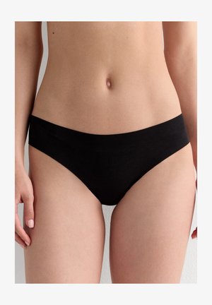 Femme portant un sous-vêtement bikini noir uni taille basse, visible de la taille jusqu’aux cuisses supérieures sur un fond neutre.