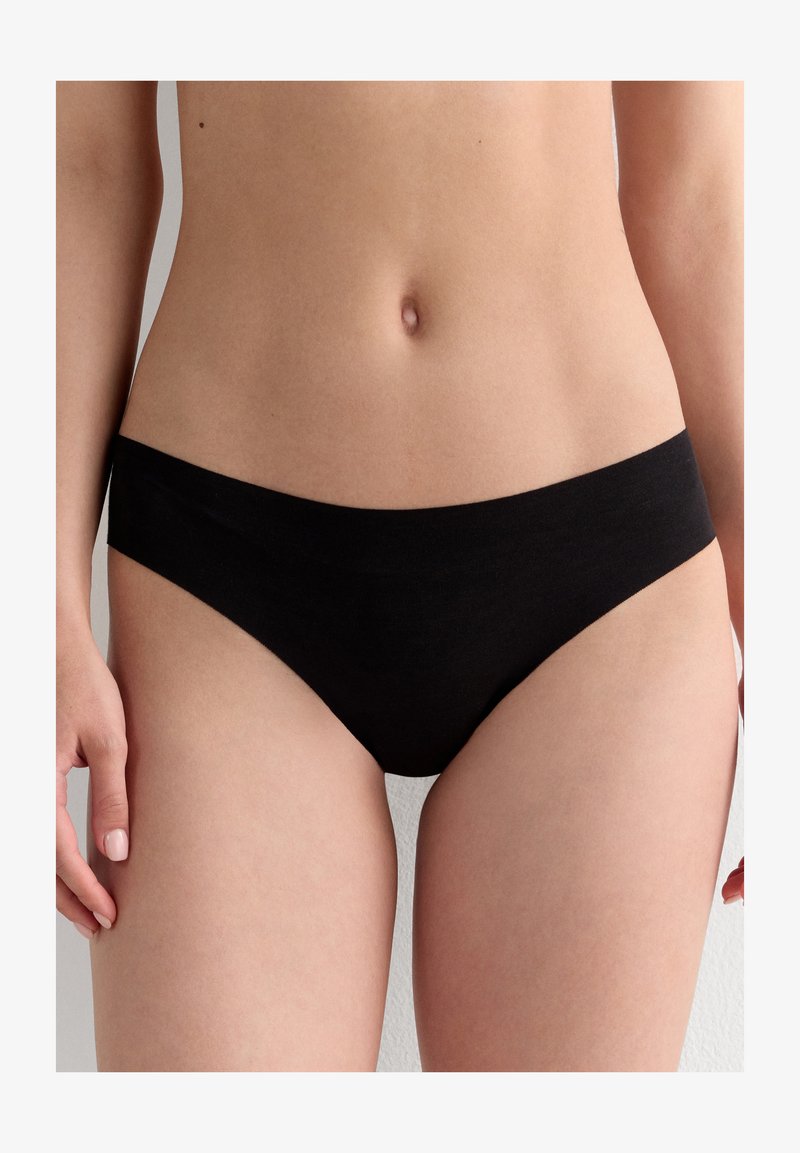 Woman wearing plain black low-rise bikini-cut underwear, shown from waist to upper thighs against neutral background.