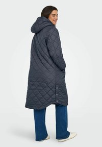 Padded tmavě šedý dlouhý péřový kabát s kapucí, bočními knoflíky a diamantovým vzorem, kombinovaný s modrými zvonovými džíny a bílými botami.