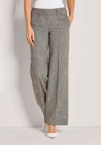 Vrouw in hooggeplaatste, wijde grijze textuurbroek en witte puntige hakken, staand met handen in de zakken.