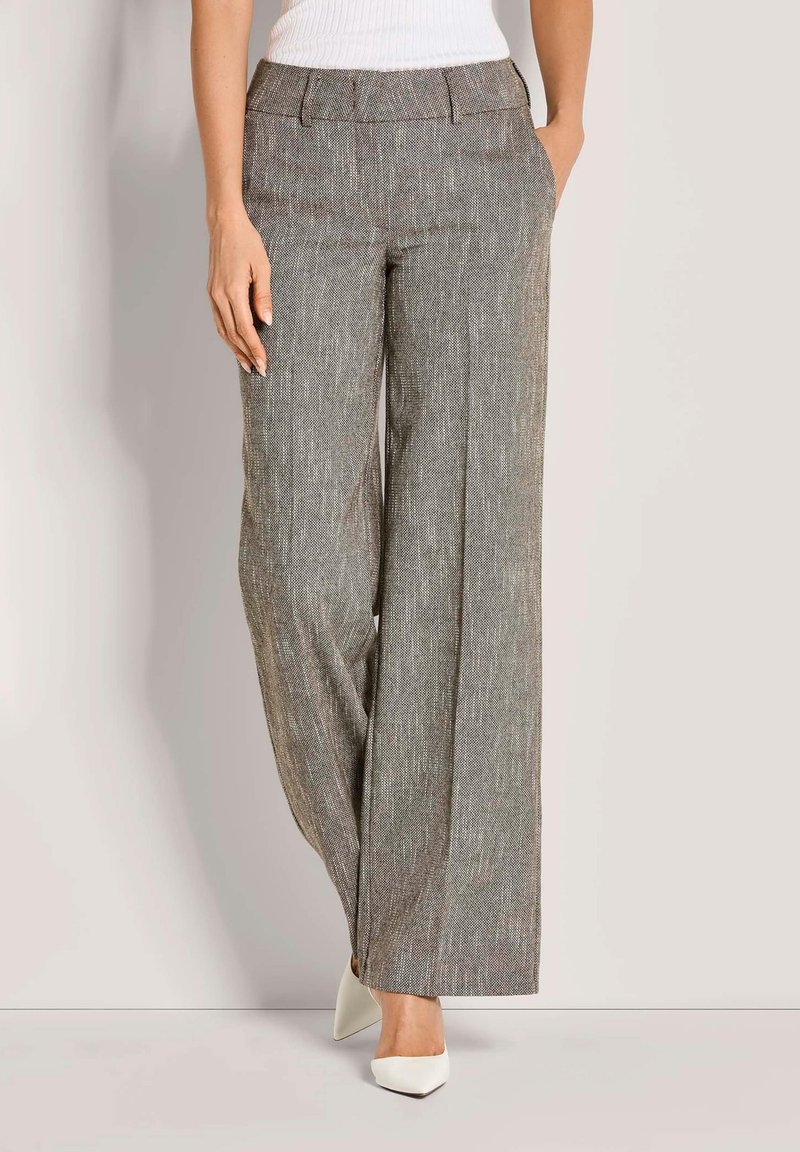 Vrouw in hooggeplaatste, wijde grijze textuurbroek en witte puntige hakken, staand met handen in de zakken.