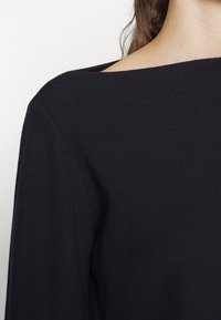 Zbliżenie osoby noszącej czarny prążkowany sweter z szerokim dekoltem, ukazujący część ramienia i szyi.