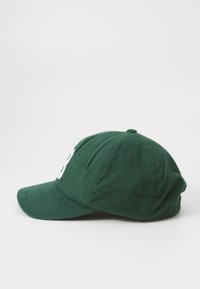 Grønn ullhatt med strukturert visir, med en fremtredende hvit logo foran og broderte detaljer langs kronen og visiret.