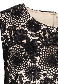Svart spetsöverdrag med blommönster på en beige tygbas. Har en rundad halsringning och en dold dragkedja på baksidan.