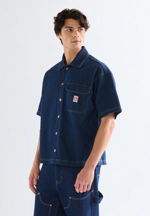 Noor mees, kes kannab tumel sinist lühikeste varrukatega teksariidest särki ja sobivaid teksapükse, vaatab veidi kõrvale tavalise heleda tausta ees.