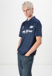 Marinblå stripete kortermet skjorte med krage, med hvite logoer og året "1990." Laget av lett stoff. Kombinert med mørke denimjeans.