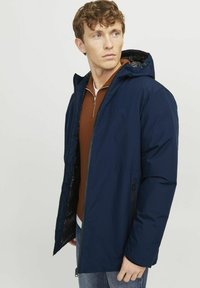 Veste isolante bleu marine avec capuche et fermeture éclair, dotée de poches latérales et d'un tissu lisse et texturé. Intérieur doublé visible.