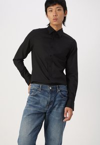 Camisa negra de manga larga con cuello puntiagudo, cierre frontal de botones y una textura suave, combinada con unos vaqueros de mezclilla azules de corte clásico.