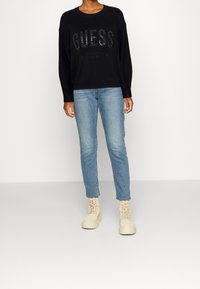 Schwarzer Pullover mit erhabenem, glitzerndem "GUESS"-Logo. Kombiniert mit hellblauen Slim-Fit-Jeans und beigen Schnürboots. Schlichtes, modernes Design.