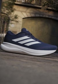 Σκούρο μπλε αθλητικό παπούτσι Adidas με λευκές ρίγες και παχύ λευκό πέλμα Cloudfoam τοποθετημένο σε πλακόστρωτο δρόμο έξω.