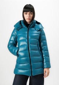 Vrouw met recht, donker haar draagt een teal gewatteerde jas met capuchon, ritssluiting aan de voorkant en zakken, poserend tegen een witte achtergrond.