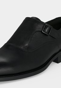 Chaussure de ville en cuir noir avec une bride à boucle et un petit talon, avec des panneaux de cuir lisses et texturés.