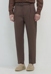 Mann in braunen Maßhosen mit Gürtelschlaufen und beigen Wildleder-Slippern, der vor einem neutralen Hintergrund steht.