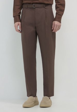 Mann in braunen Maßhosen mit Gürtelschlaufen und beigen Wildleder-Slippern, der vor einem neutralen Hintergrund steht.