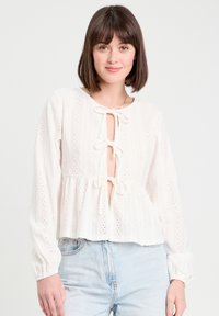 Blouse en coton blanc avec une fermeture à nouer, manches longues et style peplum. Présente des broderies œillets et des motifs texturés sur l'ensemble.