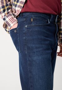 Közeli kép egy sötétkék BOSS farmernadrágot viselő személyről, akinek a keze az elülső zsebbe van téve, piros inggel és kockás zakóval párosítva.