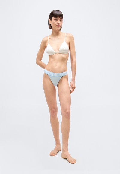 Γυναίκα όρθια, ξυπόλυτη, με λευκό μπουστάκι και ανοιχτό μπλε εσώρουχο με ψηλή γραμμή και μοτίβο στη μέση, σε απλό φόντο.
