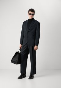 Abito nero con giacca aderente e pantaloni a gamba larga, abbinati a un dolcevita nero. Gli accessori includono occhiali da sole neri e una borsa nera con texture.