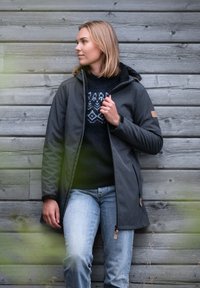 Zwarte jas met capuchon, voorzien van een rits en leren accenten, gedragen over een donkere trui met een geometrisch patroon en blauwe jeans.