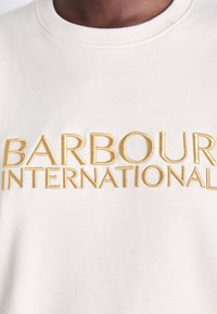 Gros plan d'un sweat-shirt de couleur crème avec le texte brodé en or "BARBOUR INTERNATIONAL" sur la poitrine.