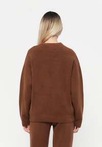 Brun stickad tröja med rund halsringning och sänkta axlar, med mjuk textur och ribbade manschetter. Sedd från baksidan.