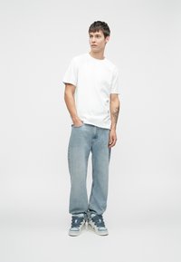 Biała koszulka w połączeniu z jasnoniebieskimi, luźnymi dżinsami i trampkami za kostkę z szarymi akcentami. Model stoi na jednolitym tle.