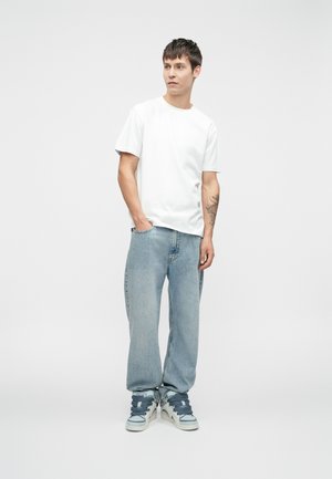 T-shirt bianca abbinata a jeans boyfriend azzurri chiari e sneakers alte con accenti grigi. Il modello si trova davanti a uno sfondo neutro.