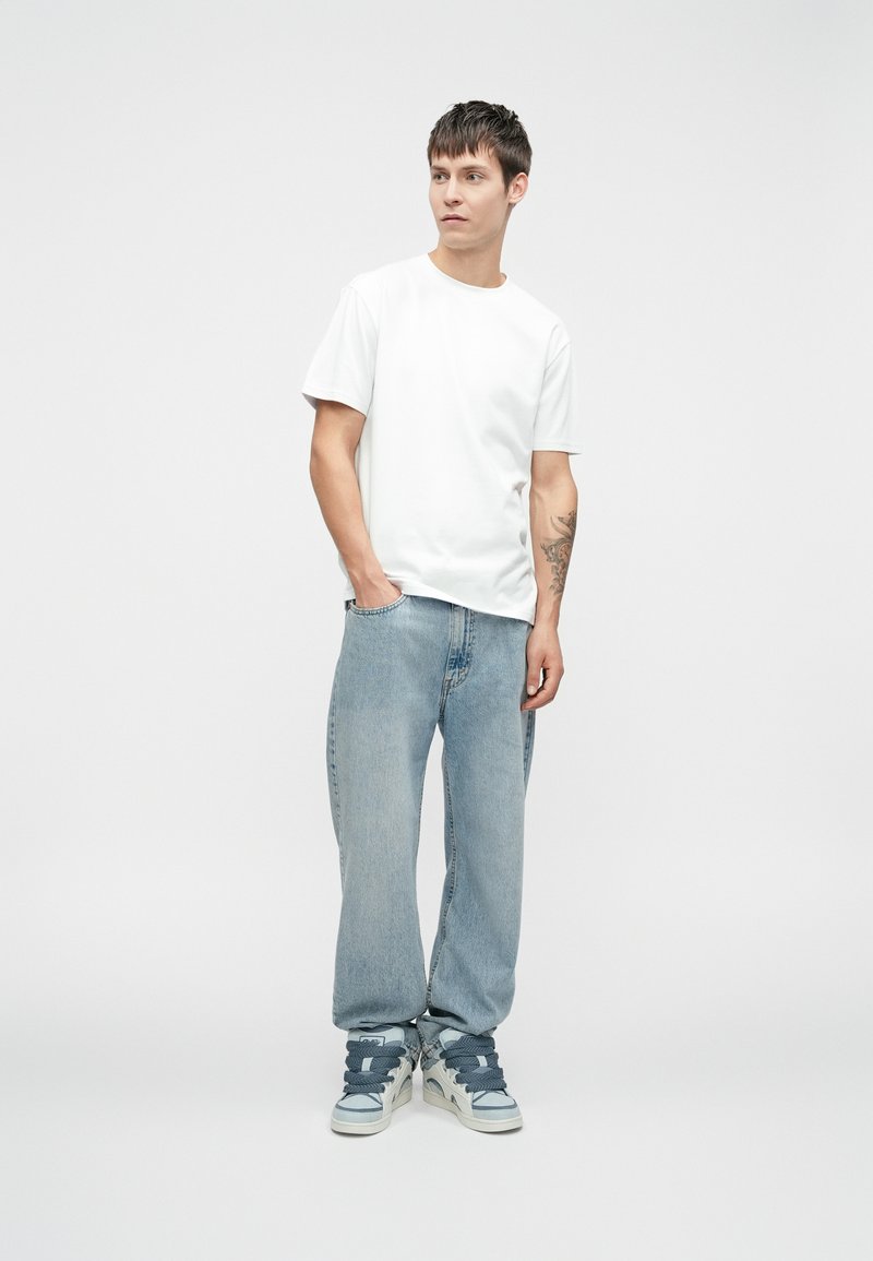 Biała koszulka w połączeniu z jasnoniebieskimi, luźnymi dżinsami i trampkami za kostkę z szarymi akcentami. Model stoi na jednolitym tle.