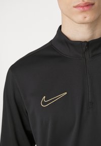 Zwarte sportshirt met rits en hoge kraag, voorzien van een gouden Nike-logo aan de bovenlinkerzijde en een gladde, rekbare stofstructuur.