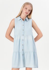 Γαλάζιο τζιν φόρεμα με γιακά πουκαμίσου, χωρίς μανίκια, κλείσιμο με κουμπιά μπροστά και φουσκωτή, κλιμακωτή φούστα. Μαλακή υφή υφάσματος.