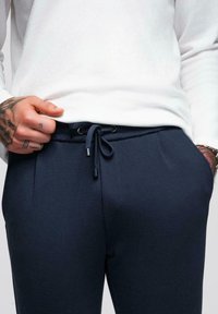 Personne portant une chemise blanche à manches longues et un pantalon bleu marine à cordon de serrage, une main tatouée ajustant la ceinture, l'autre main dans la poche.