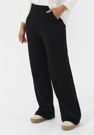 Black wide-leg pants with an elastic waistband, featuring side pockets. Worn with beige sneakers. Smooth fabric and a relaxed fit.