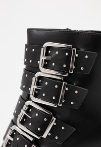 Botín negro de piel sintética hasta el tobillo con múltiples hebillas plateadas y acentos de tachuelas, con un acabado elegante y una forma estructurada.