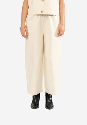 Pantalon beige à jambes larges associé à des bottines noires à bouts pointus, accessoirisé de bagues et bracelets en or aux deux mains.