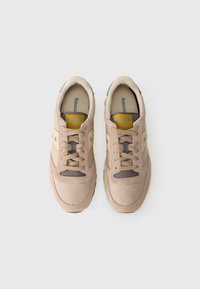 Sneakers in suede beige con punta arrotondata, dettagli in mesh e lacci tonali. Logo "Saucony JAZZ" giallo sulla linguetta. Suola in gomma piatta.