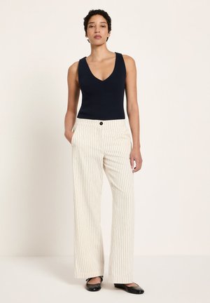 Frau steht mit den Händen in den Taschen, trägt ein ärmelloses schwarzes Oberteil, weiße, breit geschnittene Hosen mit Nadelstreifen und schwarze Ballerinas vor einfachem Hintergrund.