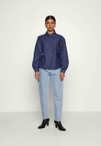 Camisa de ganga azul escura com botões e mangas longas volumosas; combinada com jeans de perna reta azul clara e botas de tornozelo pretas de salto.