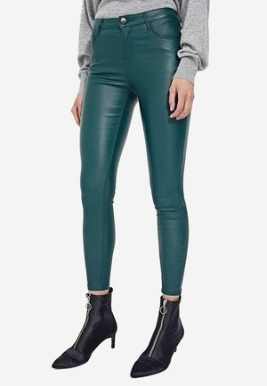 Vrouw draagt hooggetailleerde smalle teal leren broek, grijze longsleeve en zwarte enkellaarsjes met ritssluitingen aan de voorkant en kleine hakken.